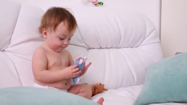 Little kid in diaper plays with toys on white sofa with pillows — Stock Video