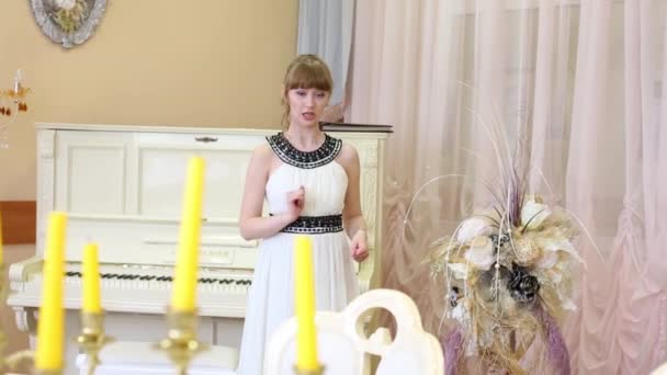 Söt flicka i vit klänning reciterar och gestikulerar nära piano — Stockvideo