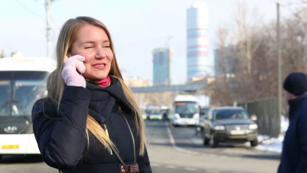 Женщина стоит рядом с дорогой в городе и говорит по телефону в зимний день — стоковое видео