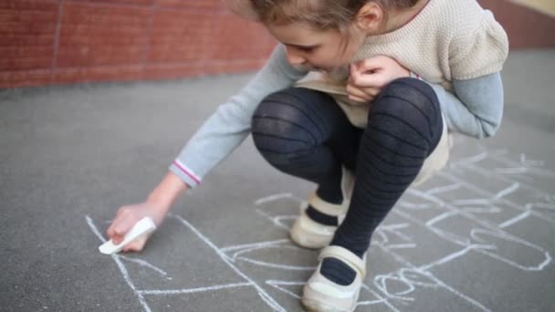 Little girl draws hopscotch on asphalt by white chalk on street — Stock Video