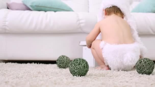 Ребенок в белой мягкой шляпе сидит на ковре с шариками возле дивана — стоковое видео