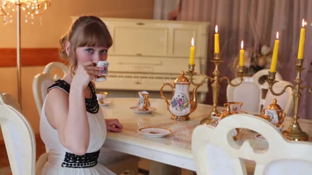 Niña se sienta a la mesa con velas encendidas y sostiene la taza de té — Vídeos de Stock