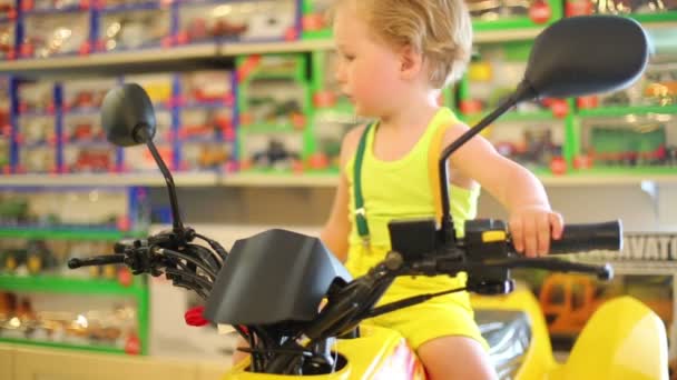 Niño en amarillo se sienta en la motocicleta de juguete en la tienda con juguetes — Vídeos de Stock