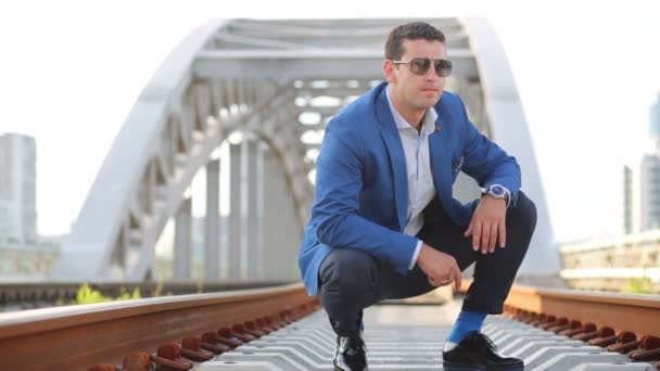 Young man in sunglasses squats and takes stones on railway bridge — Stock video