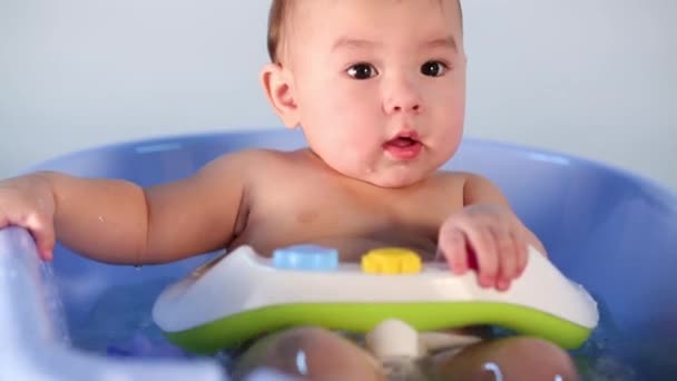 Маленький мальчик сидит в синей ванне и брызгает водой — стоковое видео