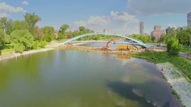 Экскаватор грузит грунт к грузовику возле моста через пруд — стоковое видео
