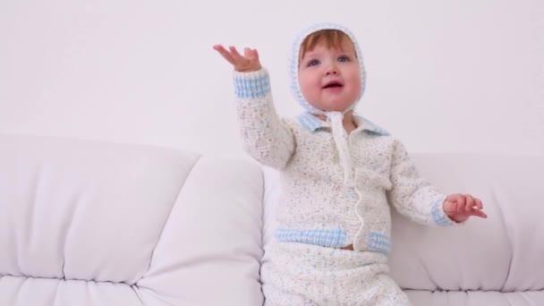 Pequeno bebê em chapéus e terno de malha senta-se no sofá branco em casa — Vídeo de Stock