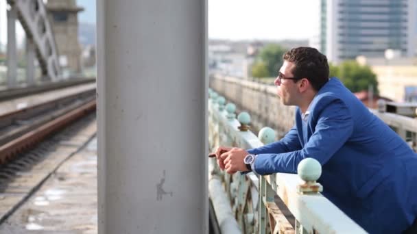 Joven con chaqueta azul fuma cigarro en puente ferroviario — Vídeos de Stock