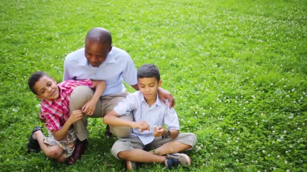 Человек, сидящий на траве в летнем парке со своими сыновьями — стоковое видео