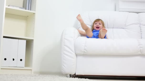 La niña se sienta en el sofá, cuelga las piernas y agita la mano — Vídeos de Stock