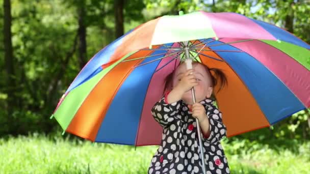 Улыбающаяся маленькая девочка с красочным зонтиком стоит на газоне — стоковое видео