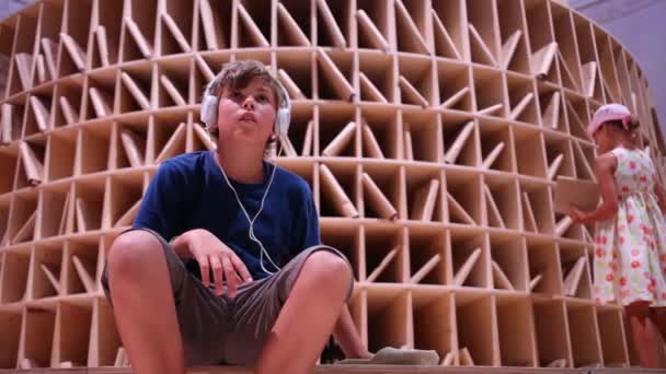 Boy with headphones sits near bookshelves — Stock Video