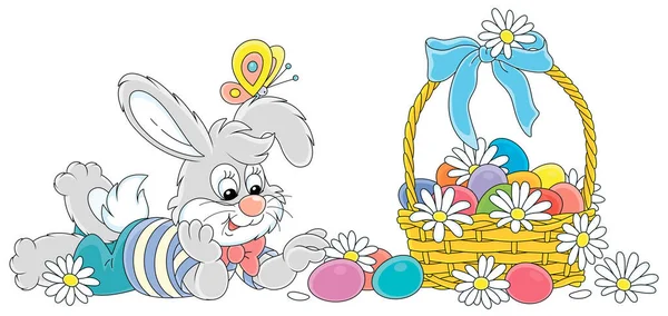 白い花と青いリボンの弓で飾られたカラフルなイースターエッグの幸せな小さな灰色のウサギとウィッカーバスケット 白い背景にベクトル漫画のイラスト — ストックベクタ