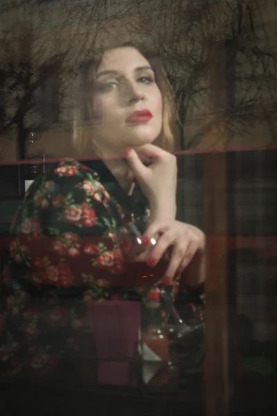Hermosa mujer se sienta en un café, sosteniendo una taza, fotos detrás de gla — Foto de Stock