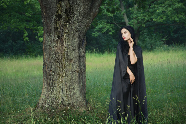 Beautiful brunette woman in black dress and black cloak in the magic forest. Gothic style.