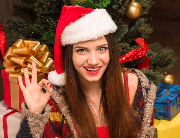 Красивая девушка сидит перед новогодней елкой и показывает хорошо знак — стоковое фото