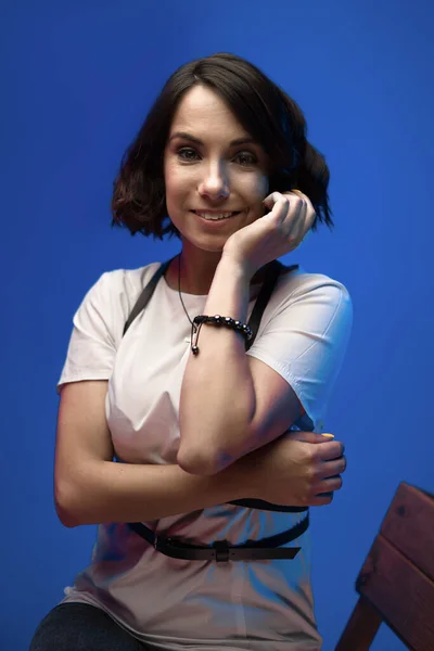 Wanita tersenyum mengenakan t-shirt putih menyentuh pipinya dengan tangan berpose di latar belakang biru di studio — Stok Foto