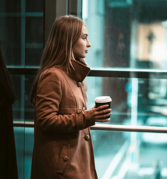 İş merkezinde kahve içen genç bir kadın. Uzun saçlı, ceketli Avrupalı bir kadın elinde camdan dışarı bakan bir kağıt bardak tutuyor. — Stok fotoğraf