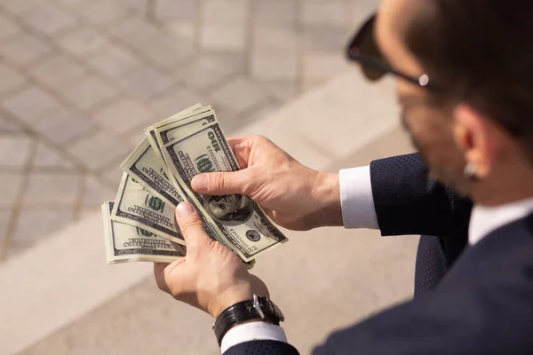 Close up of successful american businessman considering wad of 100 dollar banknotes while sitting on sidewalk. Rear view — Stock Photo, Image