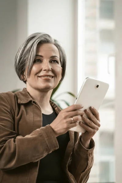 Pretty woman of retired age smiles communicating online on digital tablet while standing near window. Technology concept — Stock Photo, Image