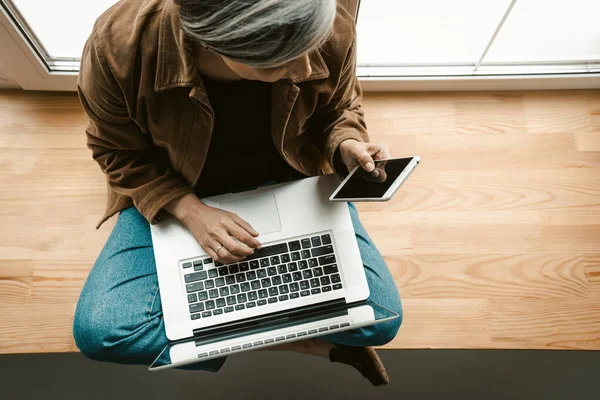 Drukke vrouw kijkt op het mobiele scherm werken laptop computer terwijl zitten op grote houten vensterbank. Bovenaanzicht. Kopieer ruimte rechts — Stockfoto