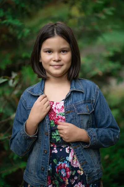 Nettes kleines Mädchen in kariertem Kleid und Jeansjacke posiert vor dem Hintergrund der grünen Natur — Stockfoto