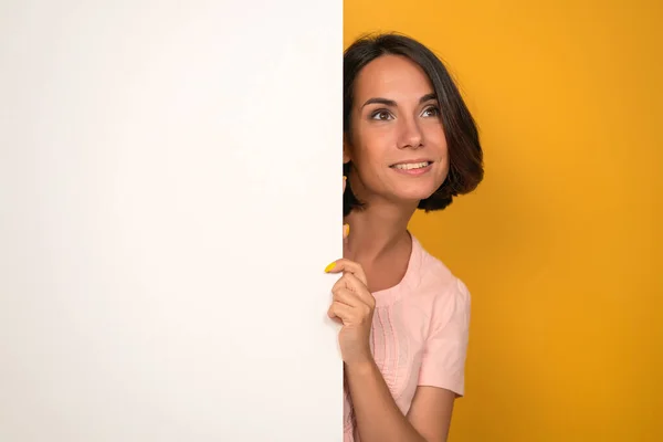 Молодая женщина смотрит вверх, держа белый пустой лист с копировальным местом на нем. Изолирован на жёлтом фоне. Снимок студии — стоковое фото