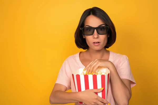 Siyah gözlüklü, elinde patlamış mısır kutusu olan genç bir kadın. Sinema konsepti. Sarı arka planda izole edilmiş. Boşluğu sola kopyala — Stok fotoğraf