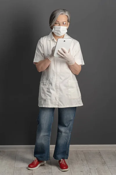 Médica feminina vestindo casaco branco e jeans de comprimento total. Mulher madura em máscara protetora trabalhando com dispositivo digital — Fotografia de Stock