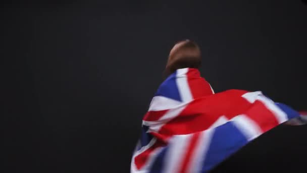 Malá blondýnka se točí a končí na anglické vlajce. Bělošské dítě se usmívá na vlajku na ramenou na černém pozadí. — Stock video