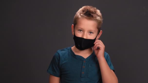 A kisfiú leveszi a fekete maszkot a kezével a fekete háttéren. A kaukázusi gyerek megkönnyebbülten lélegzik és mosolyog a kamerába nézve. A világjárvány megszüntetésének fogalma. — Stock videók