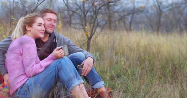 Mladý pár sedící na podložce si užívá a odpočívá venku. Bělošský mladík hladí svou přítelkyni. Usměje se a drží hrnek v ruce. — Stock video