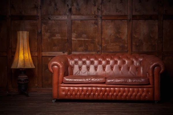 Hermoso sofá de cuero chester marrón. En el interior del loft de color oscuro, con una pared de madera en el fondo. Hay dos interiores: un sofá y una lámpara de pie. —  Fotos de Stock