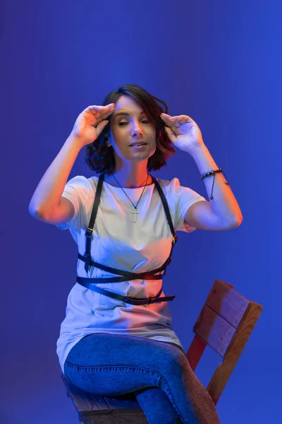 Mujer joven de cabello oscuro con el pelo corto se sienta en la silla con las manos arriba alisando su cabello. Alegre dama caucásica aislada sobre fondo azul. —  Fotos de Stock