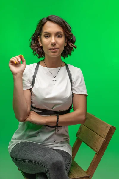 Jolie jeune femme en t-shirt blanc assise sur une chaise. Dame gaie avec cheveux courts foncés découpés sur fond vert chromatique. — Photo