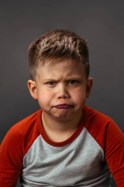 Liten pojke är stygg och grimaser med missnöje. Det missnöjda barnet visar sina känslor. Studio porträtt på grå bakgrund — Stockfoto