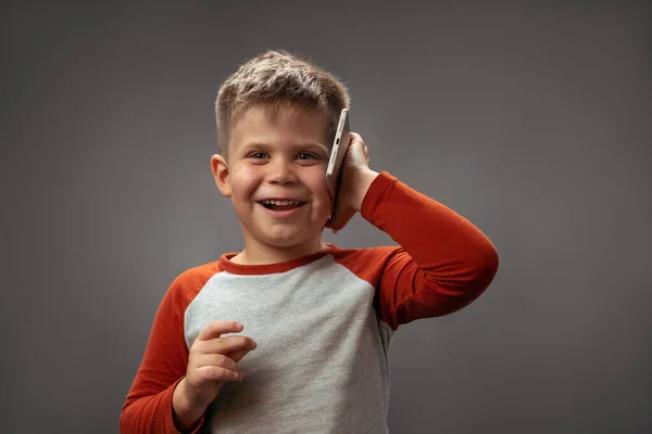 En liten kaukasisk pojke med kort frisyr. Glädjande kommunicerar i telefon. På en grå bakgrund — Stockfoto