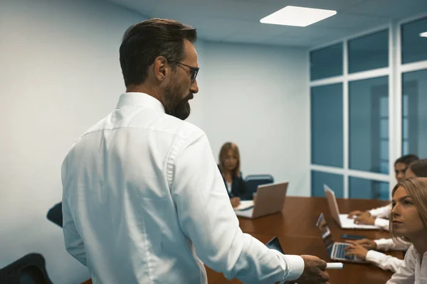 オープンスペースのオフィスで大きなテーブルで働いているチームとの議論を持っている前に彼の後ろに立ってビジネスマン — ストック写真