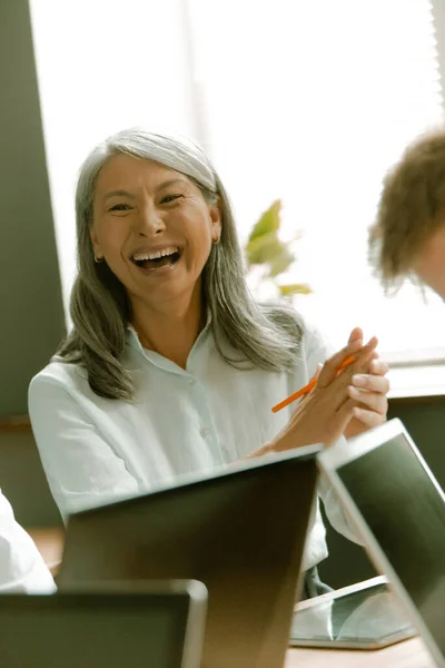Divertente asiatica anziana donna ride di riunione d'affari seduta alla scrivania dell'ufficio con computer portatili su di esso contro di finestra sullo sfondo — Foto Stock