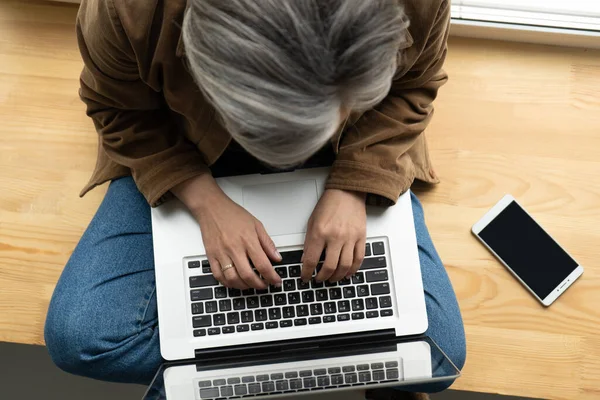 Een grijze vrouw die op de computer werkt. Bovenaanzicht van Kaukasische blogger of freelancer met behulp van internet op laptop zittend op houten vensterbank met mobiele telefoon erop. close-up schot — Stockfoto