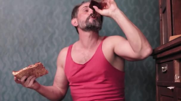 Un bărbat de vârstă mijlocie cu barbă bea dintr-o sticlă şi miroase o plăcintă. Poartă un tricou roşu. Conceptul alcoolismului masculin. Imagini FullHD de înaltă calitate — Videoclip de stoc
