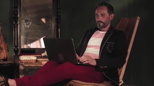 一位中年男子坐在笔记本电脑后面，坐在椅子上工作。背景音乐风格别致。笔记本电脑在它的膝盖上 — 图库视频影像