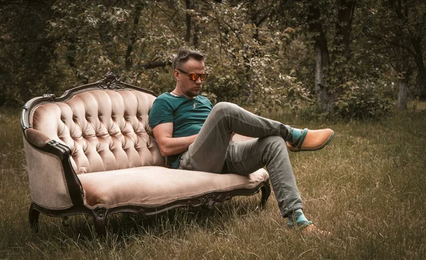 Отдыхающий мужчина в серых джинсах сидит на диване посреди сада — стоковое фото