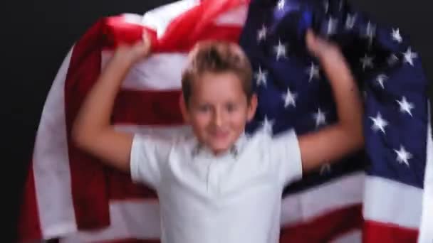 Kleine jongen zwaait Amerikaanse vlag achter zijn rug tegen zwarte achtergrond. Kaukasische 10-jarige kind verheugt zich en glimlacht. — Stockvideo