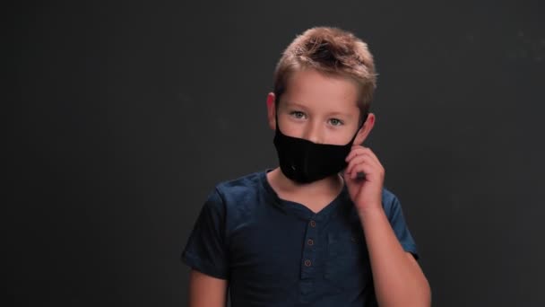 Küçük çocuk siyah arka planda siyah maskesini çıkartıyor. Beyaz çocuk rahatça nefes alıyor ve kameraya bakıp gülümsüyor. Salgını sona erdirme kavramı. — Stok video