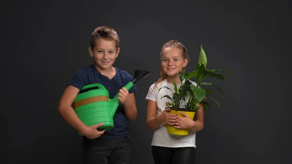 Adolescentes caucasianos rindo, um menino segurando uma lata de rega, uma menina segurando uma planta em um vaso de flores. Isolado em fundo cinza escuro. Conceito de cuidado vegetal — Fotografia de Stock