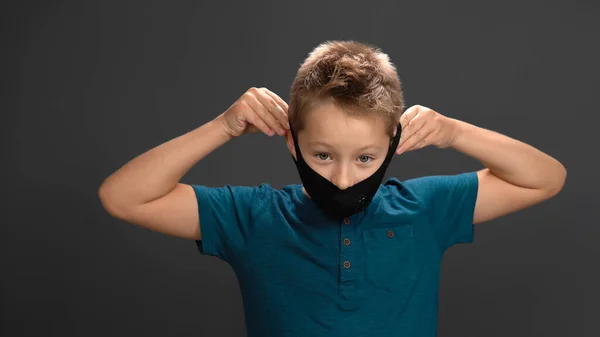 Den lille pojken tar av sig sin svarta mask med handen på grå bakgrund. Kaukasiska barn andas med lättnad och ler tittar på kameran. Begreppet utrotningspandemi — Stockfoto