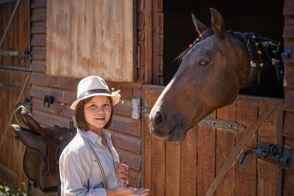 Niña jinete se comunica con el caballo después del deporte ecuestre. La yegua marrón se asoma fuera del establo de manera amistosa. Temas animales — Foto de Stock