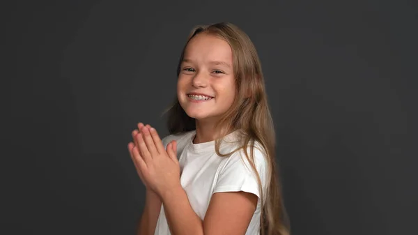 Heureuse jeune fille aux cheveux longs sourit largement applaudissements mains. Belle écolière caucasienne avec système de bretelles sur les dents. Isolé sur fond gris. Espace de copie — Photo