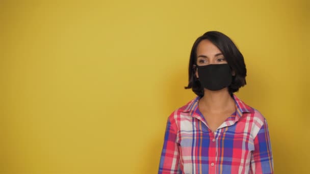 Mujer joven de pelo corto con cabello oscuro sobre fondo amarillo con máscara negra en la cara mira cuidadosamente a un lado. Modelo en ropa de todos los días vale la pena. El concepto es virus, pandemia. Copiar espacio. — Vídeos de Stock